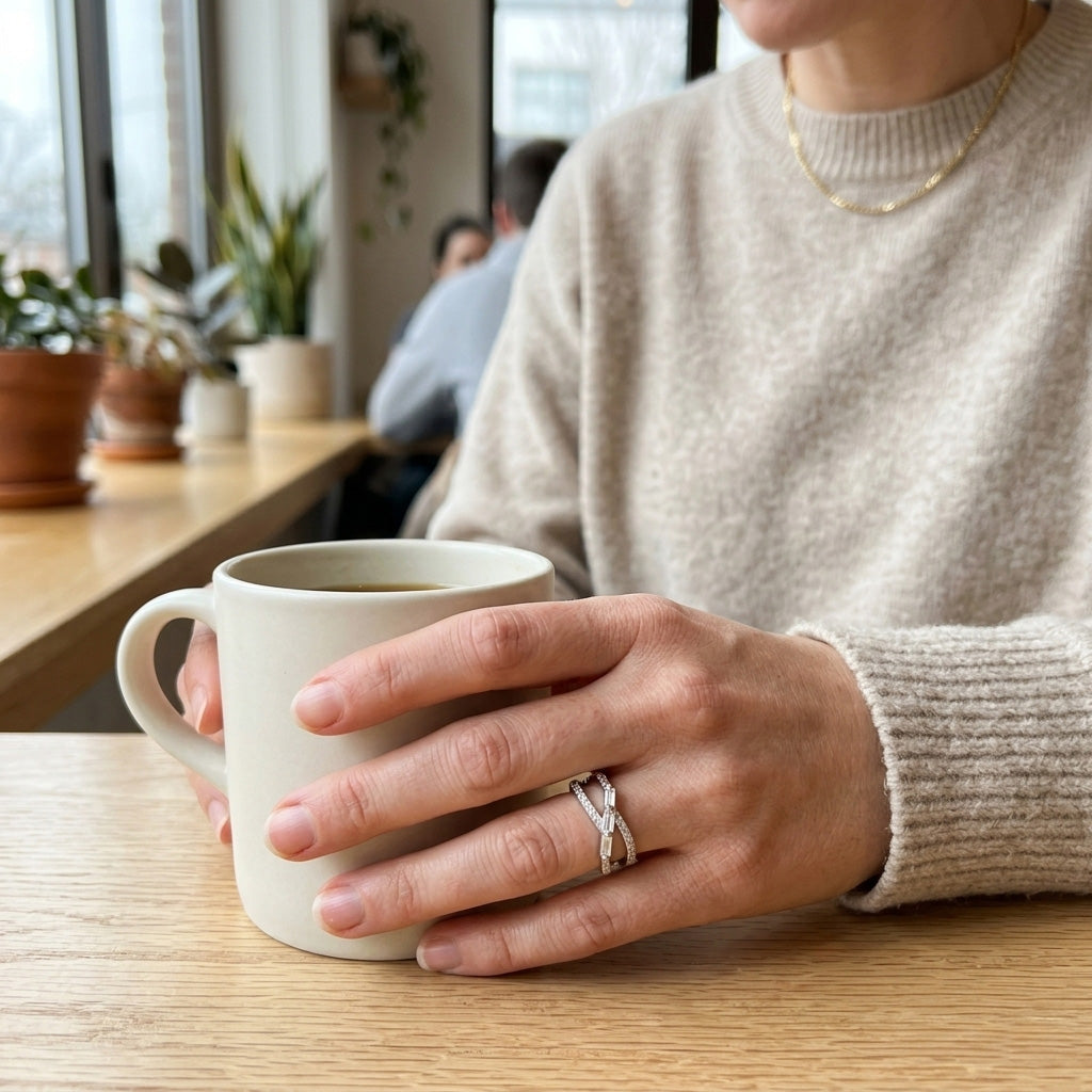 Lifestyle shot of a woman in a beige sweater holding a white coffee mug, showcasing a Triple Baguette Crystal Crossover Silver Ring for daily wear.