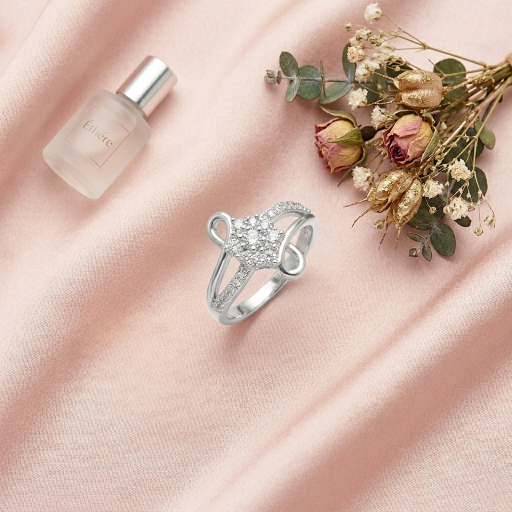 Flat lay of the Silver Infinity Swirl Crystal Ring on pink silk next to a small frosted perfume bottle and dried miniature roses.