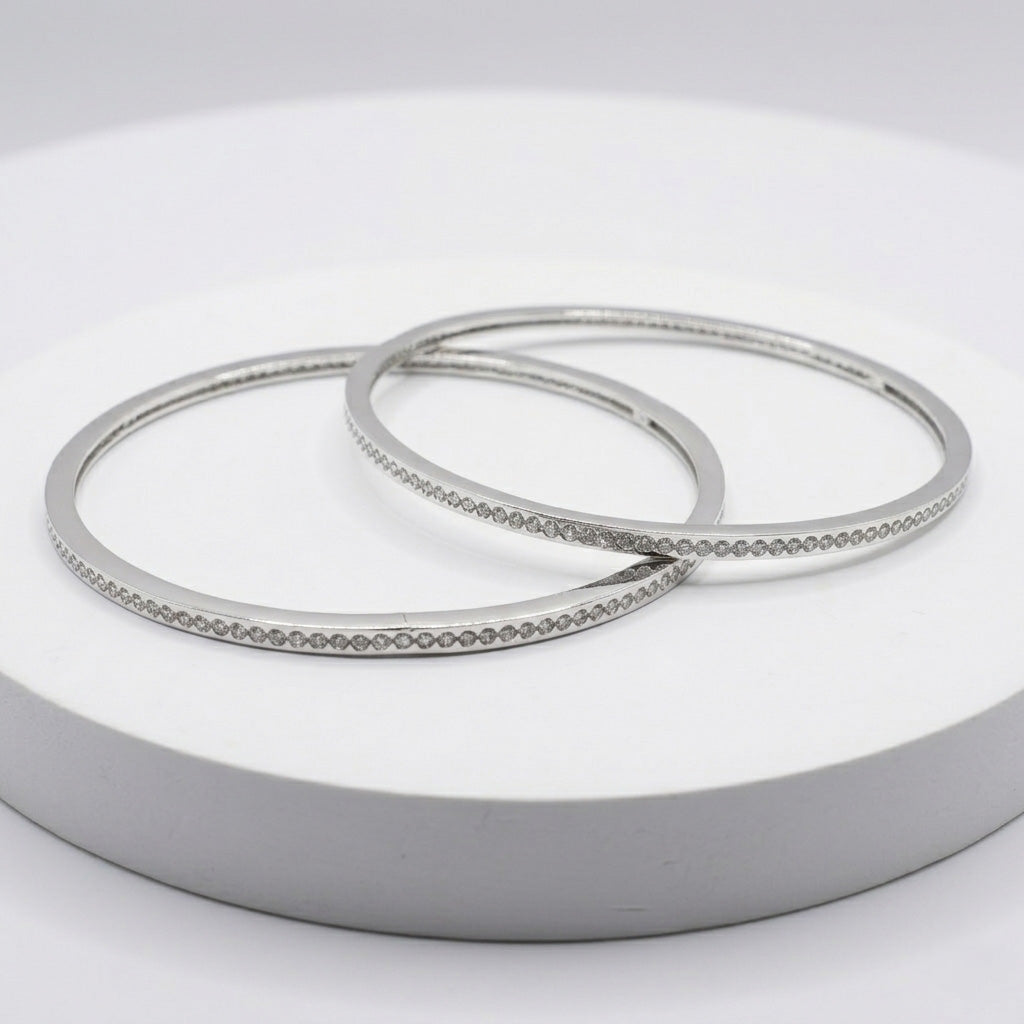 The Slim Stone Silver Bangle Pair displayed flat on a minimalist white circular podium, highlighting the sleek, continuous loop design and full-circle recessed stone setting
