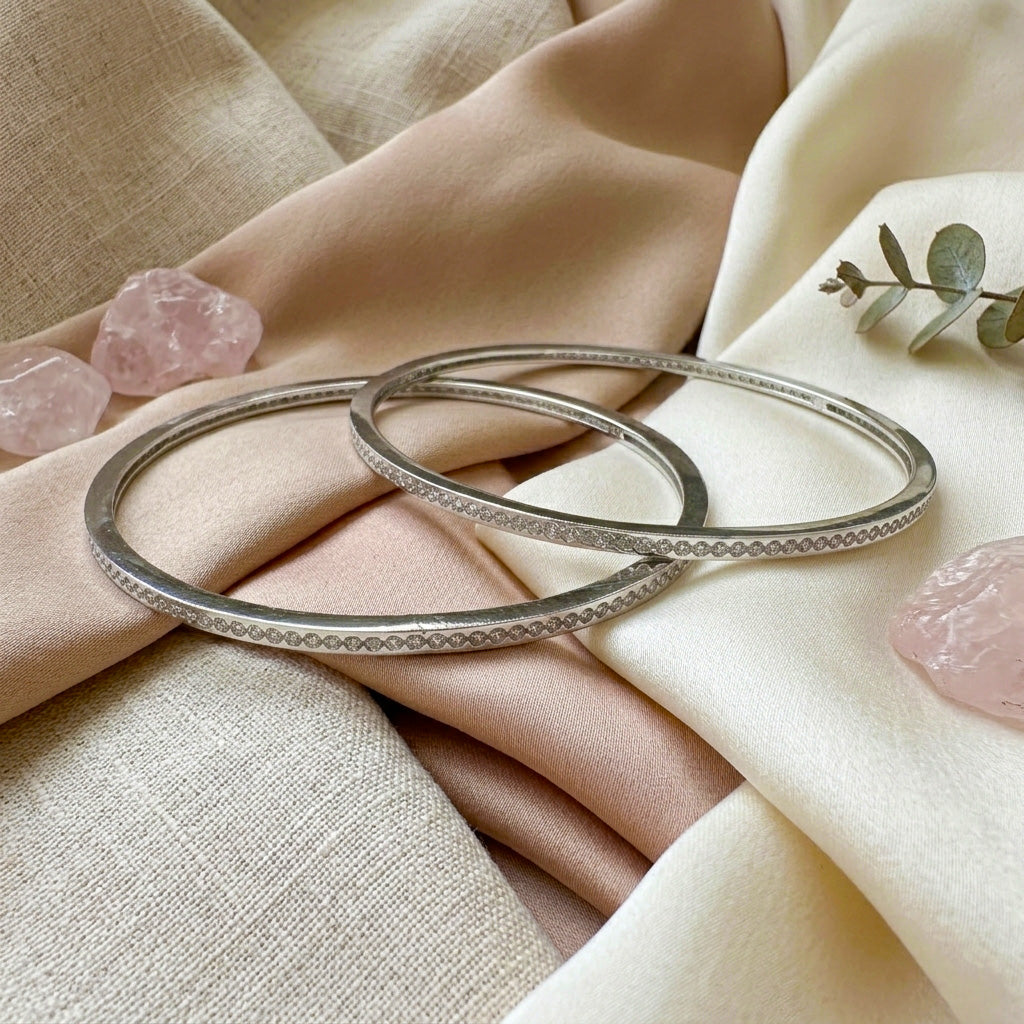 A lifestyle shot of the Slim Stone Silver Bangle Pair resting aesthetically on draped peach and cream fabrics