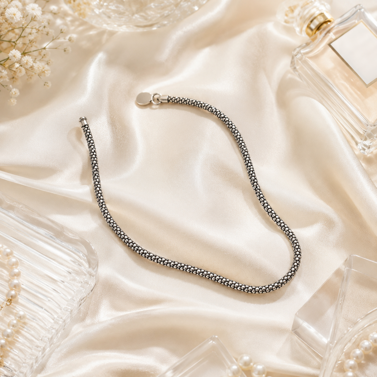Textured mesh silver anklet laid flat, revealing the detail of the signature round metal tag near the fastening.