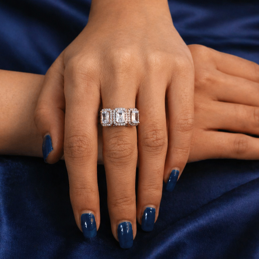 A woman's hand displaying the Emerald Cut Three-Stone Halo Ring against a dark blue velvet background.