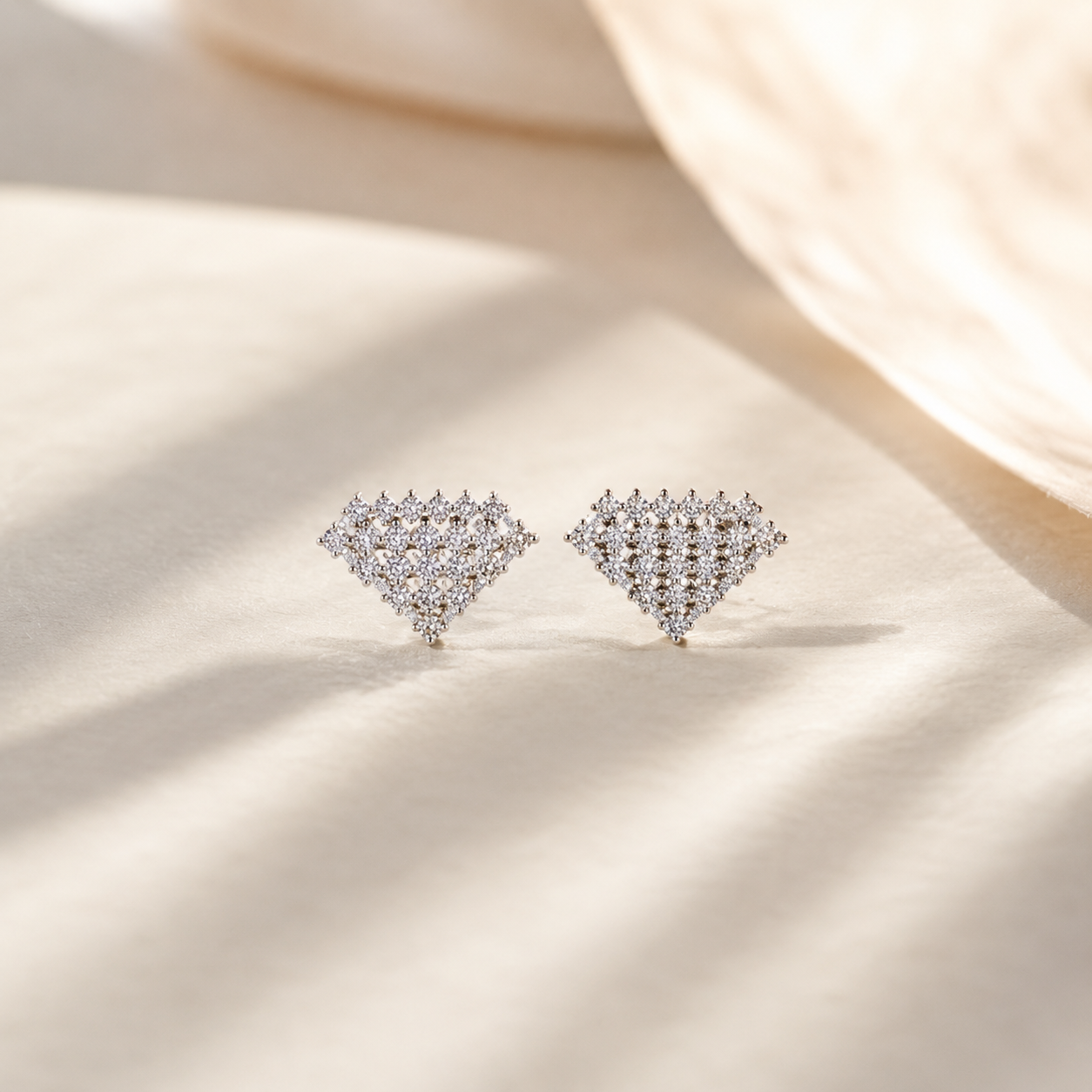 Close-up of Geometric Diamond-Shaped Silver Crystal Stud Earrings resting elegantly on a textured cream surface with soft warm sunlight. 