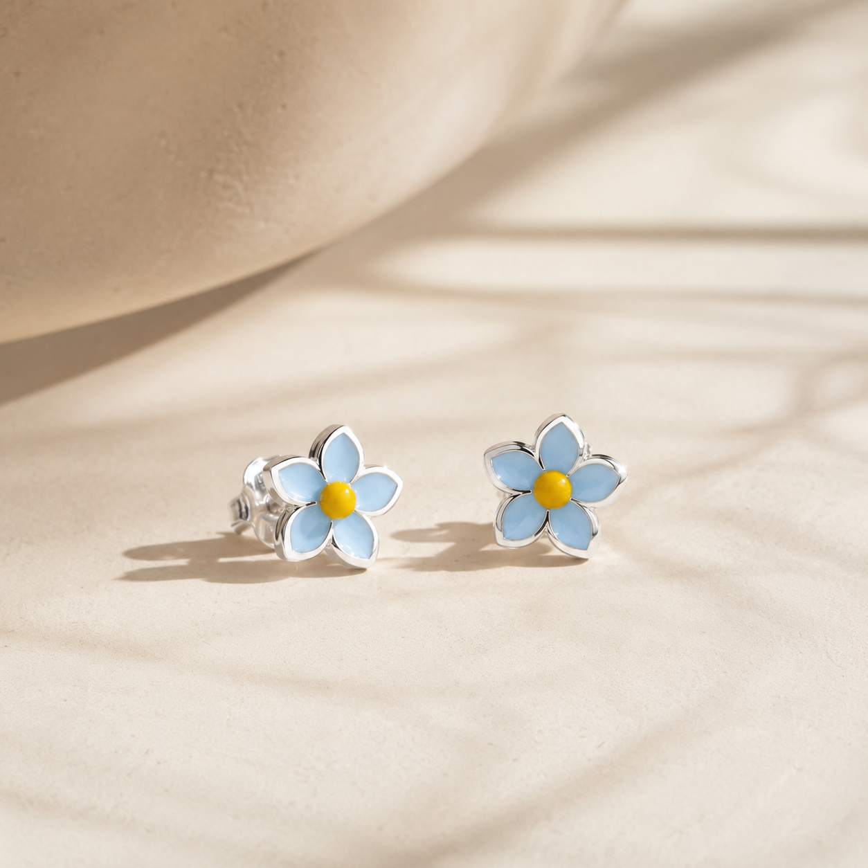 Pair of Light Blue Floral Enamel Silver Stud Earrings resting gracefully on a textured beige surface with soft sunlight shadows.