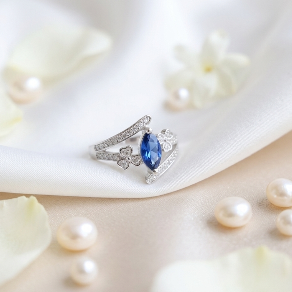 Close-up of the Neelam Blossom Silver Bypass Ring resting on white silk fabric surrounded by pearls and soft white flower petals.