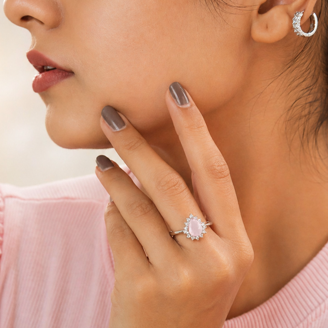Close-up of a woman touching her cheek, elegantly displaying the Rose Quartz Pear Halo Silver Ring on her hand 