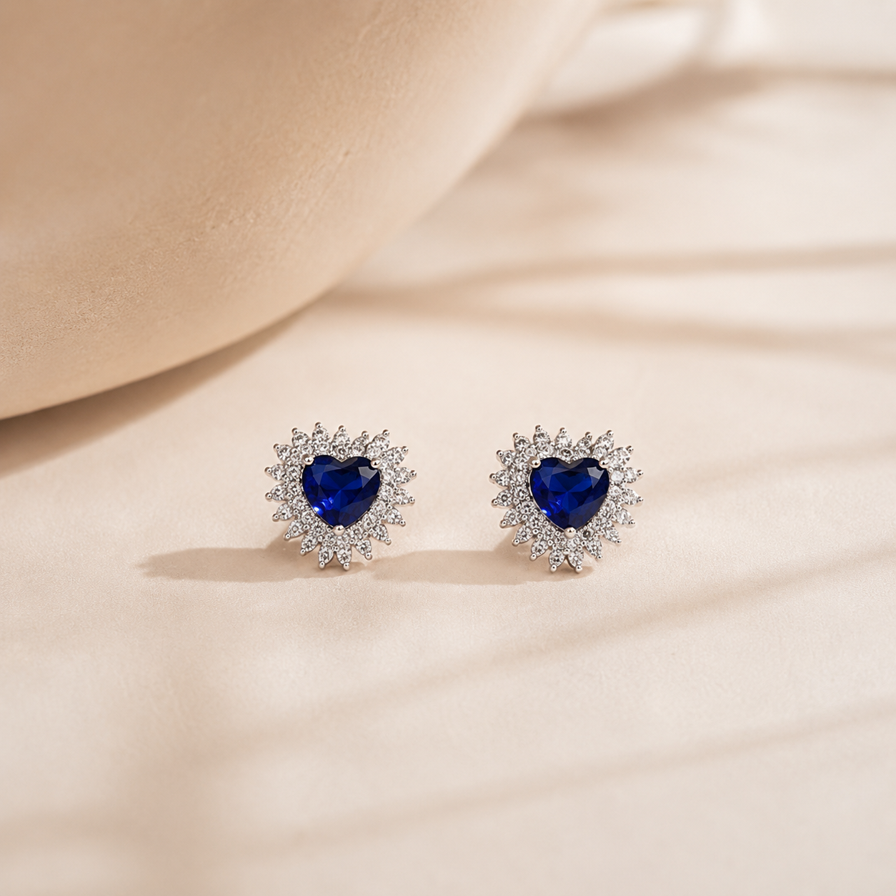 Pair of Royal Blue Heart Sunburst Halo Stud Earrings resting on a smooth, neutral textured surface with elegant shadow play