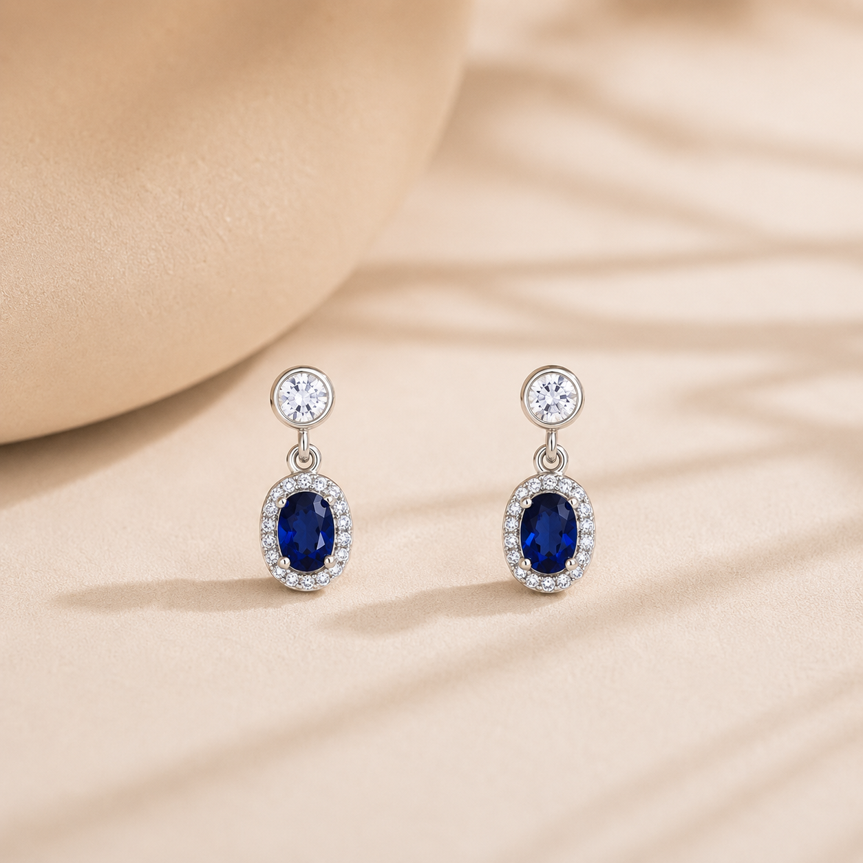 Sapphire Blue Oval Halo Drop Earrings are beautifully displayed on a warm beige textured surface with soft shadow patterns.