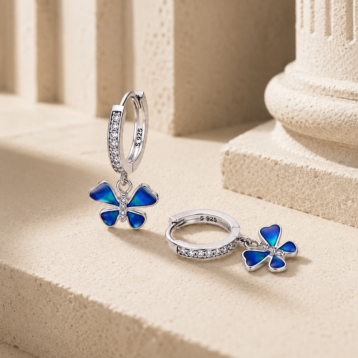 Pair of Silver Blue Enamel Petite Butterfly Huggie Drop Earrings displayed on a stone surface, showing the enamel wings and CZ detailing