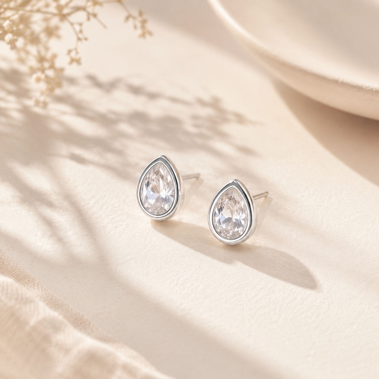 Pair of Silver Dainty Pear Cut Solitaire CZ Stud Earrings displayed on a light fabric surface, showing the pear-cut CZ and polished silver border