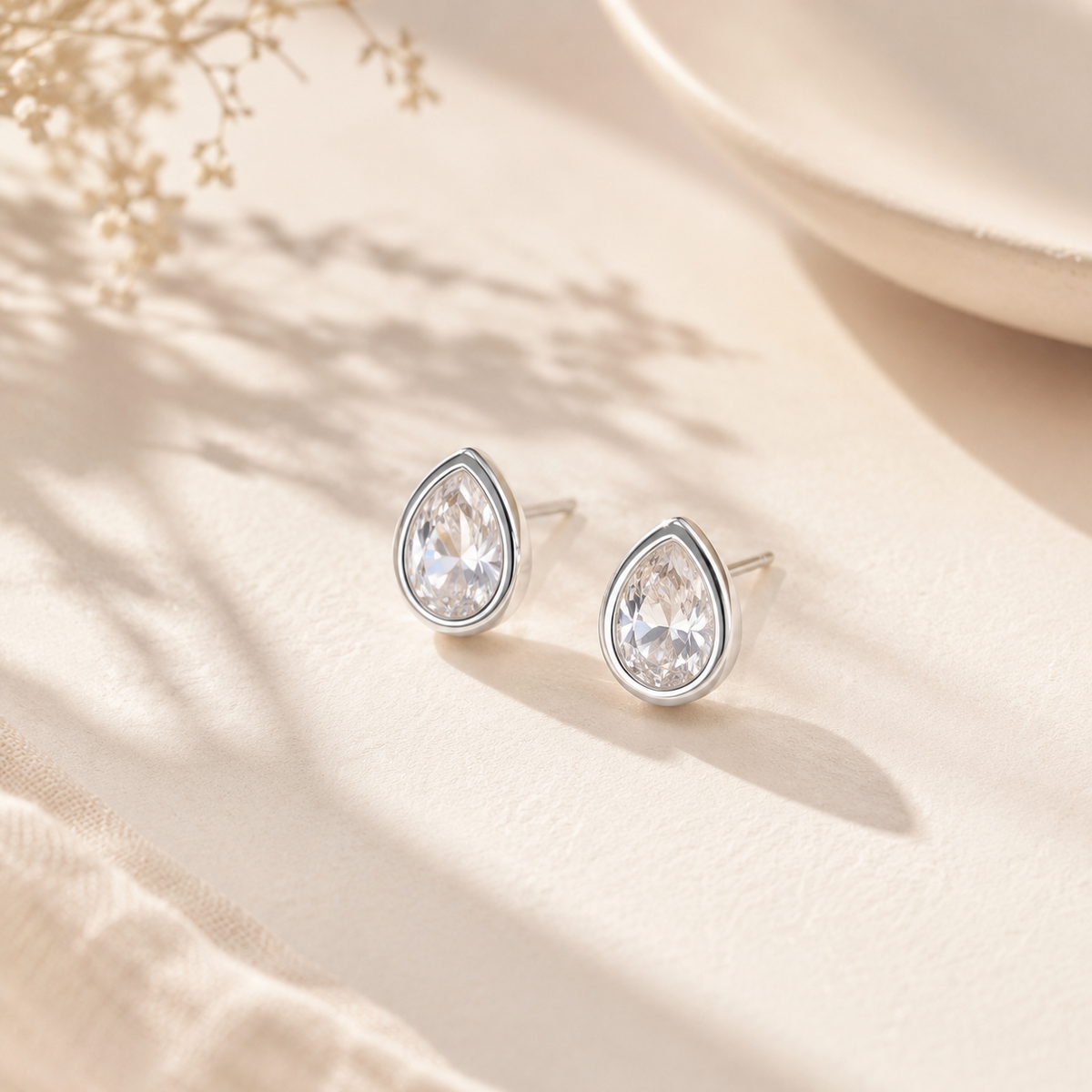 Pair of Silver Dainty Pear Cut Solitaire CZ Stud Earrings displayed on a light fabric surface, showing the pear-cut CZ and polished silver border