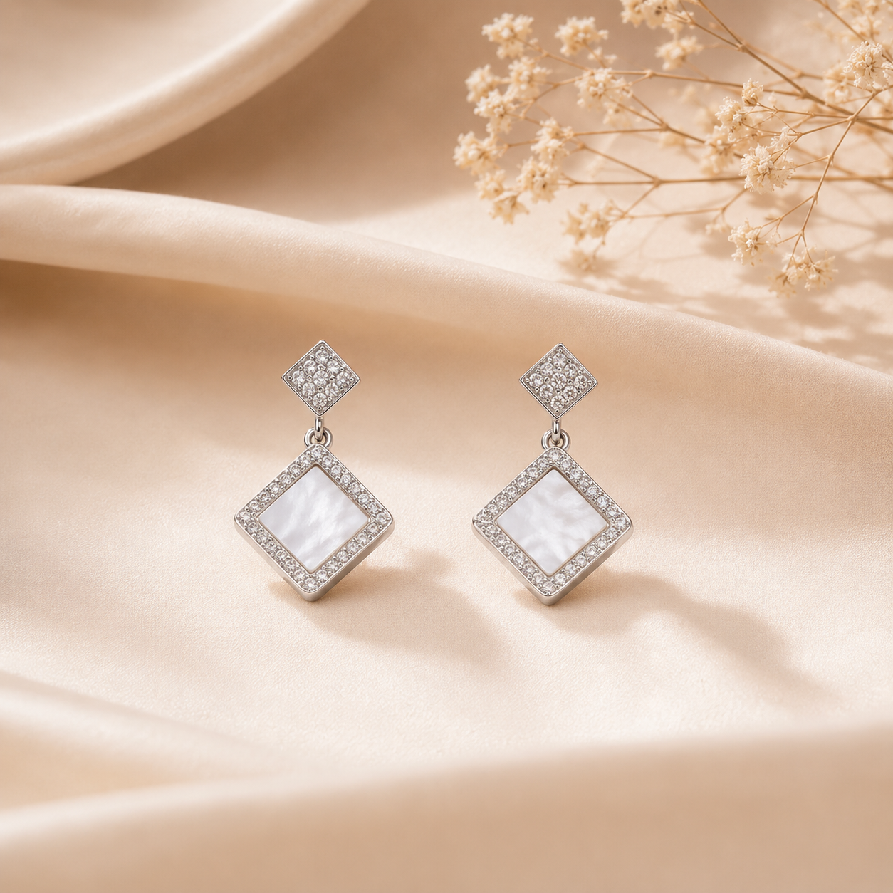 Silver Rhombus Mother of Pearl Halo Drop Earrings displayed on a soft beige silk fabric with dried flowers.