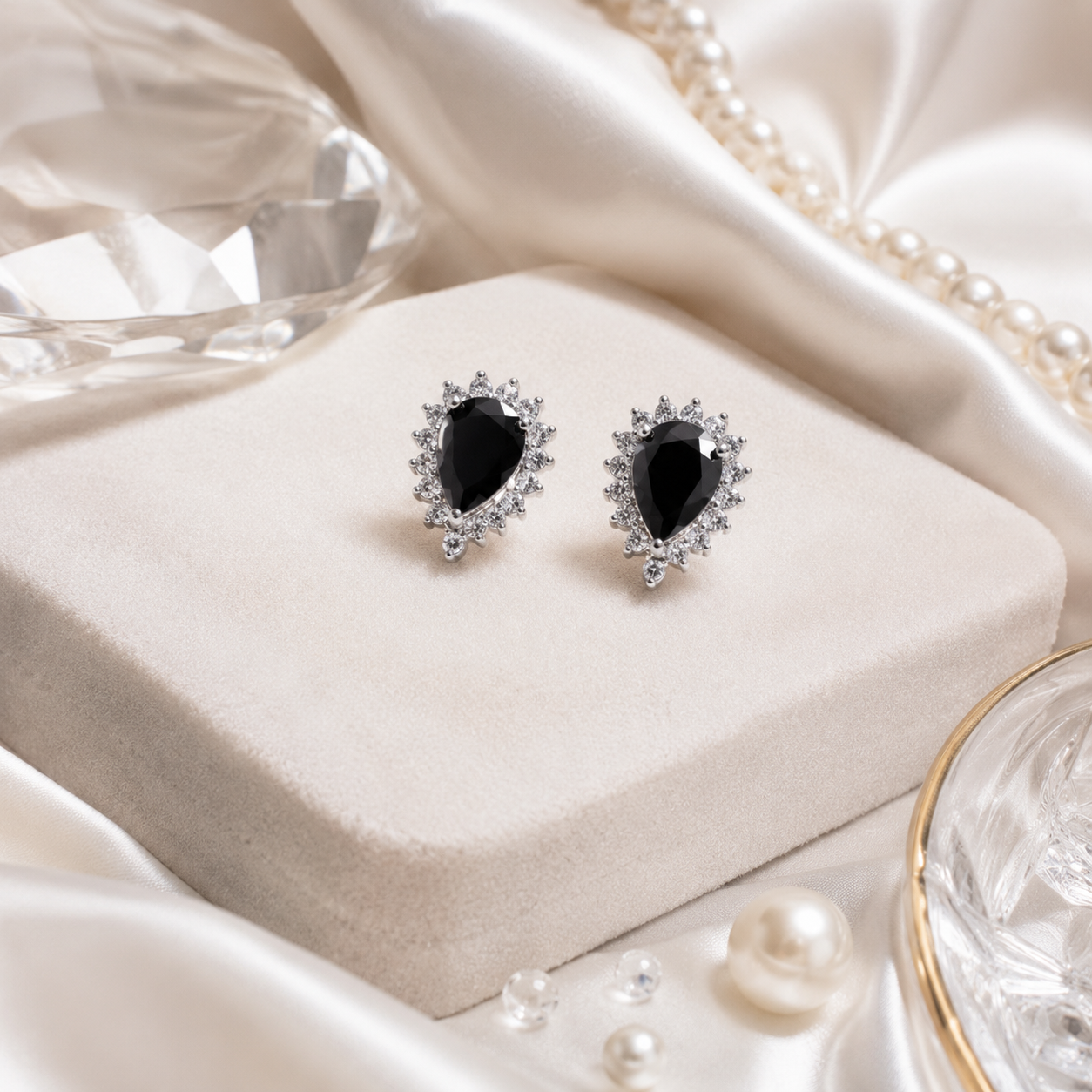 Teardrop Black Stone Halo Silver Stud Earrings are displayed elegantly on a soft beige velvet block alongside scattered pearls and a crystal prop.