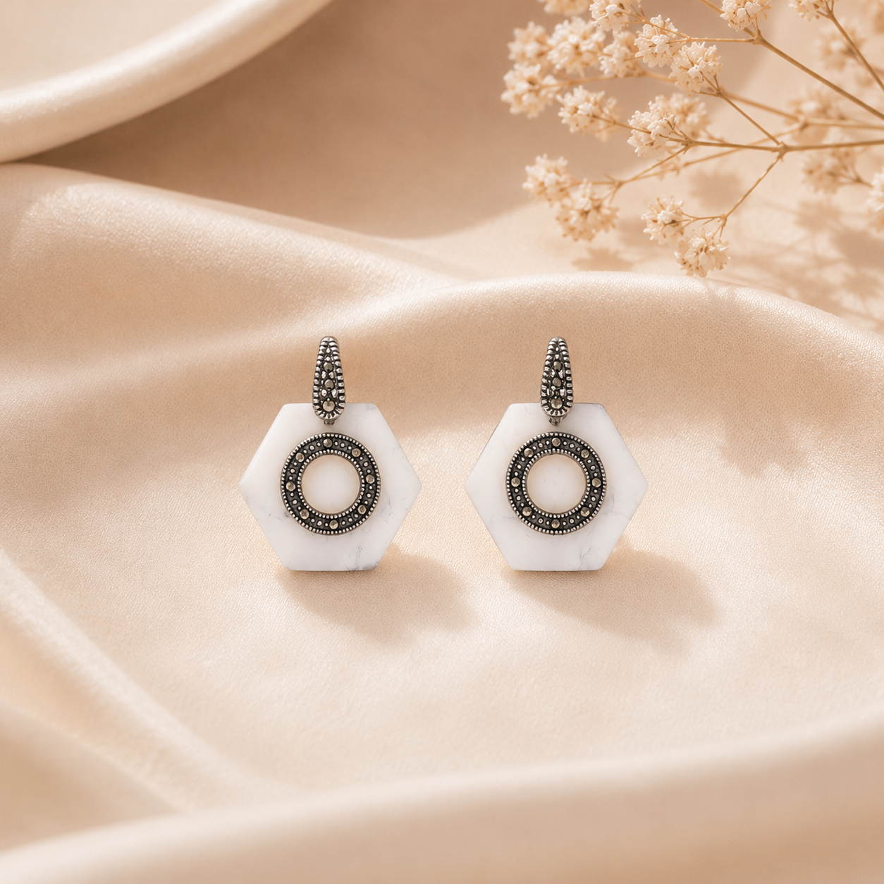 White Hexagon Oxidized Silver Statement Earrings are placed elegantly on a textured beige silk fabric next to dried baby's breath flowers.